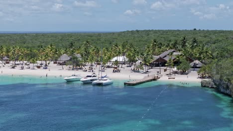 Filmische-Luftaufnahme-Mit-Korallenriff,-Klarem-Wasser,-Yachten,-Sandstrand-Mit-Touristen-Und-Grüner-Tropischer-Landschaft-An-Sonnigen-Tagen---Insel-Catalina,-Dominikanische-Republik