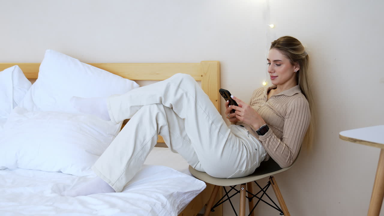 Good-looking Caucasian woman sits on the chair in the bedroom. Relaxed lady uses her smartphone surfing internet.