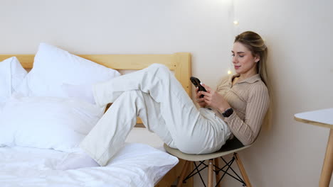 good-looking caucasian woman sits on the chair in the bedroom. relaxed lady uses her smartphone surfing internet.