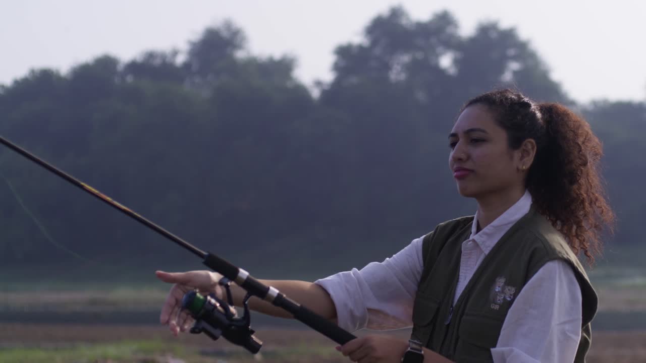 Una donna sudasiatica che tiene una canna da pesca e osserva con calma un tranquillo lago, riflettendo pazienza, concentrazione e una serena luce diurna all'aperto.