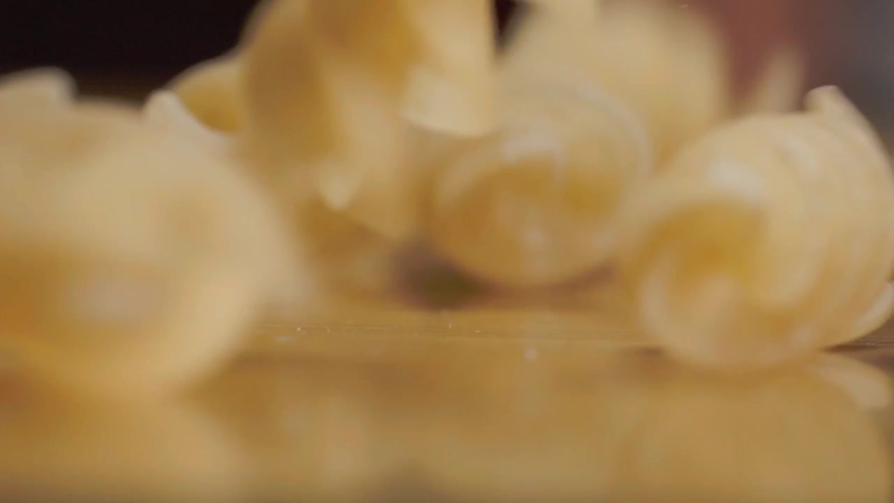 Pezzi di pasta cruda cadono su una superficie riflettente, creando movimento e riflessi tenui, mentre la messa a fuoco macro evidenzia il loro colore dorato pallido e le forme irregolari.