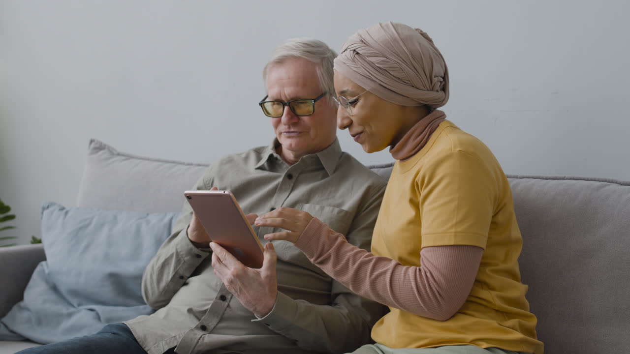 Wanita Arab Mengajari Pria Lansia Menggunakan Tablet Saat Mereka Duduk di Sofa di Rumah