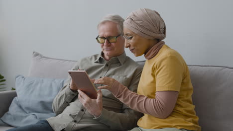 arabic woman teaching an elderly man to use tablet while they are sitting on sofa at home 1