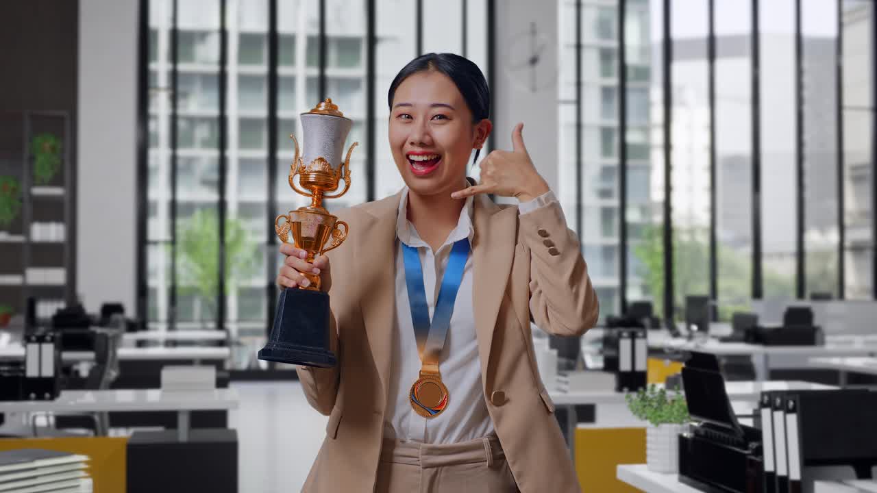 Asian Business Woman In A Suit With A Gold Medal And Trophy Showing Call Me Gesture And Smiling To Camera As The First Winner In The Office