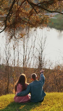 romantic couple enjoys the nature. young man and woman sitting on grass on the hill and looking at beautiful river in springtime. back view. vertical video