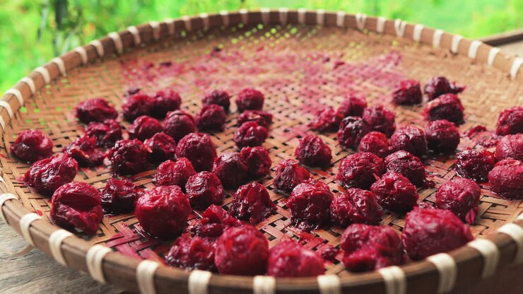 Closeup of ruby red plums drying on a woven bamboo tray in Moc Chau, Vietnam, with soft green garden bokeh behind, highlighting rustic handmade fruit preservation