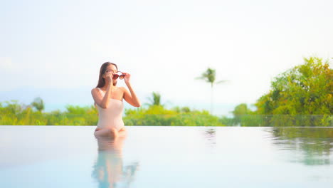 Eine-Hübsche-Junge-Frau-In-Einem-Sexy-One-Shoulder-Badeanzug-Sitzt-Am-Rand-Eines-Infinity-Pools-Und-Setzt-Ihre-Sonnenbrille-Auf