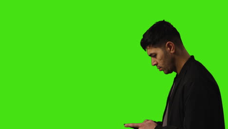 Serious-Young-Man-In-Thought-Walking-Into-Frame-Text-Messaging-On-Mobile-Phone-Against-Green-Screen-Background