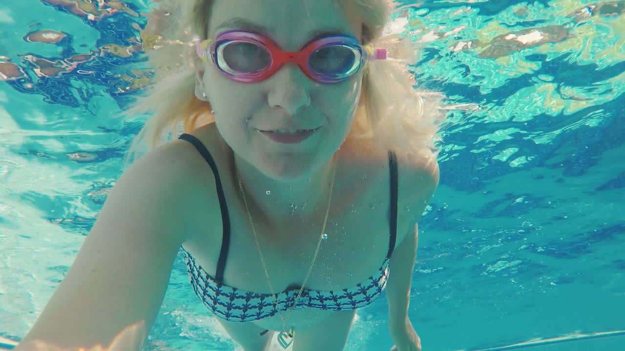 한 젊은 여성이 물속에서 비디오에서 자신을 촬영합니다. 수영장에서 수레가 카메라를 들여다 봅니다. w