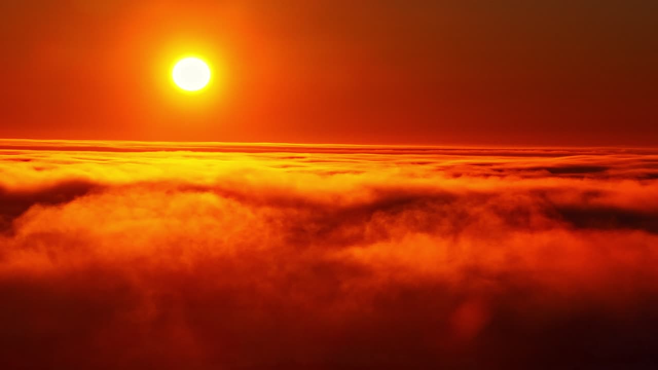 Toma de dron a gran altitud de un atardecer ardiente de tonos naranjas y rojos con el sol brillante asomándose entre nubes en rápido movimiento.