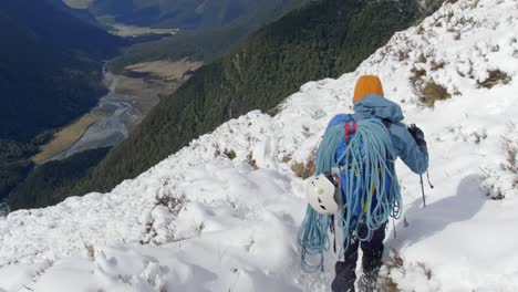 Man-climbing-mountains-walking-through-the-snow-above-a-beautiful-valley-on-a-sunny-day,-handheld-shot