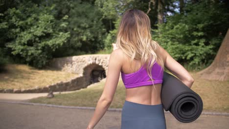 thin, fit blonde woman adjusts her hair as she walks toward a stone arch bridge in a park carrying a yoga mat