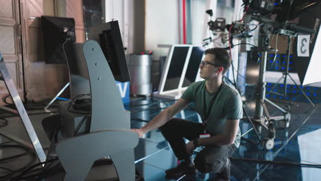 man adjusting digital equipment and monitor settings in modern studio, crouching near high-tech gear, production in process, focused on timer and screen, professional filming environment