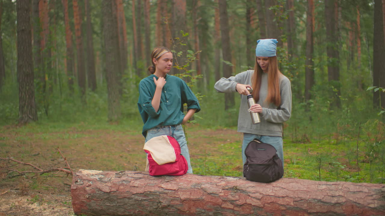 пешеходы останавливаются в спокойном лесу с рюкзаками, опирающимися на упавшее дерево, одна в синей бандане и сером свитере открывает термос, чтобы налить напиток, в то время как ее спутник в зеленой рубашке смотрит на нее