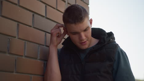 young man standing beside brick wall reaches to remove cigarette from ear while preparing to light it up for smoking, wearing black vest and dark shirt under daylight with soft urban backdrop