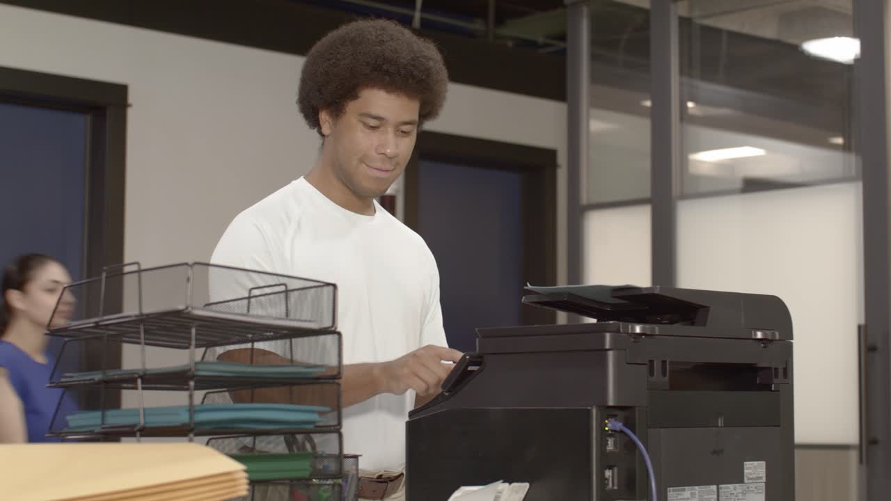 African American man at a printer in a busy office with video dolly left to right