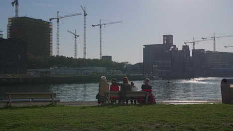 a group of people sitting on a bench in front of a river, cyclist passing by