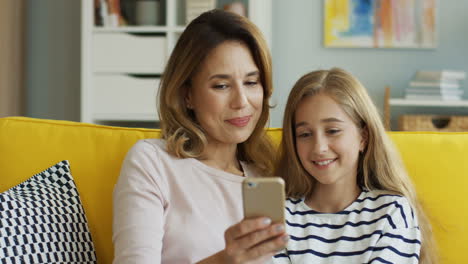 blonde mother with her teen daughter watching video on smartphone, talking and smiling while they are sitting on sofa in living room