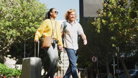 Happy-diverse-couple-walking-with-luggage-in-sunny-city-street