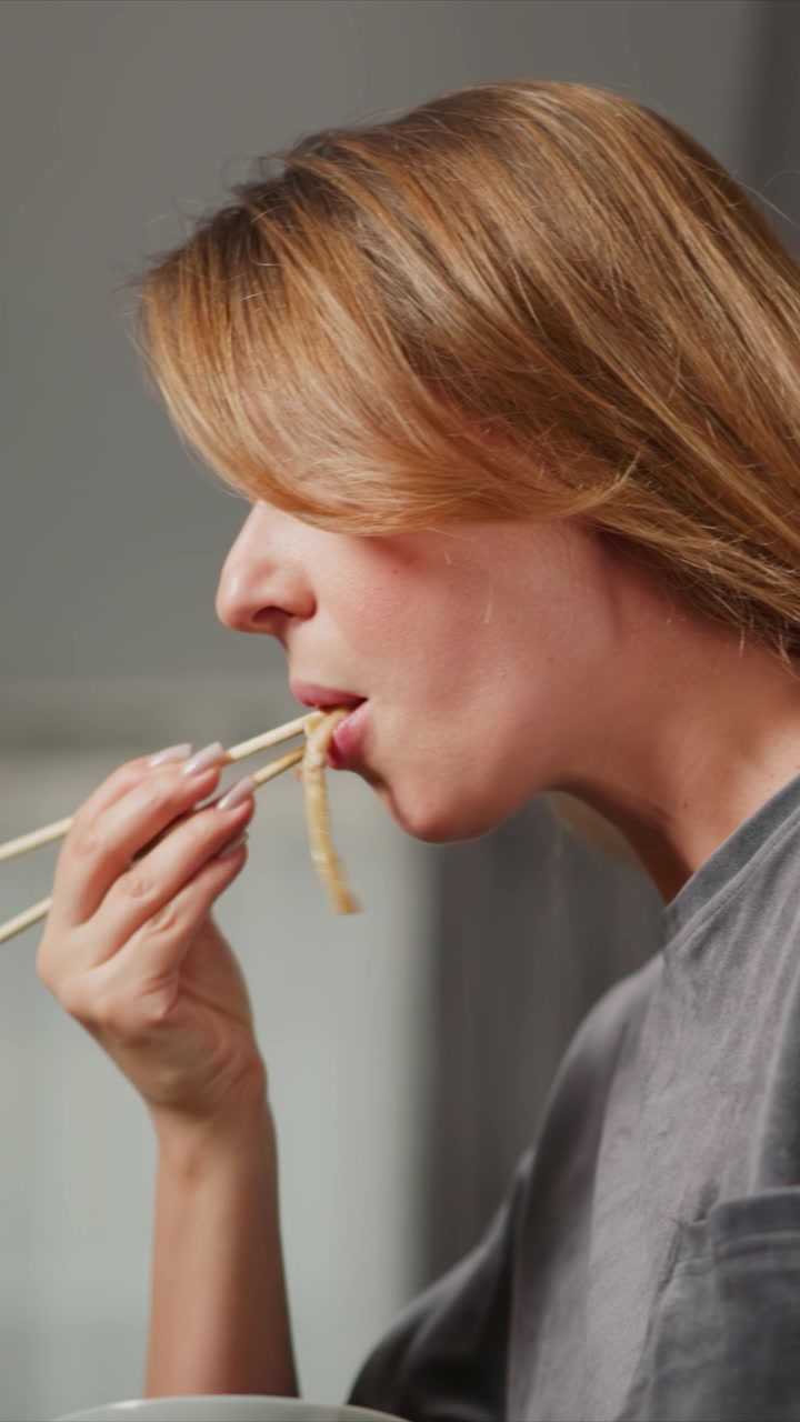 Primo piano di donna che usa le bacchette per mangiare noodles da una ciotola. La scena mostra la progressione del sollevamento e del consumo del cibo con gli utensili, concentrandosi sui movimenti delle mani e della bocca su sfondo neutro.