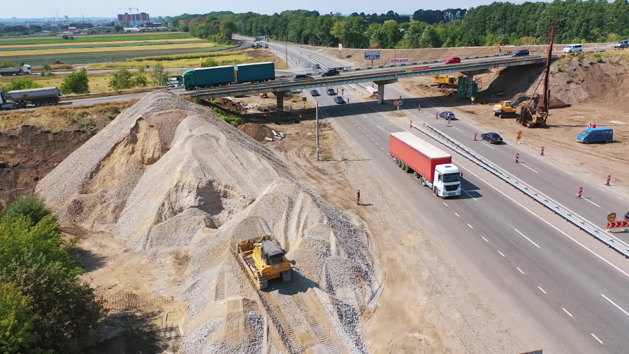 Trucks are working near the highway. Cars moving on the road. New bridge over the road with trucks and cars moving on it. Machinery for making new road in summer. Aerial view.