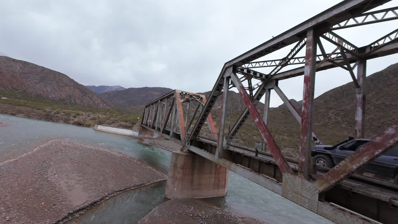 FPV drone shot of a historic steel railway bridge in the Andes, Mendoza, Argentina, part of Ferrocarril Transandino line, then a SUV enters the structure, framed by rugged terrain and river below