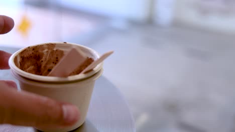 A hand stacks an empty ice cream cup into another on a round table, with soft daylight and shallow depth of field, emphasizing casual disposal