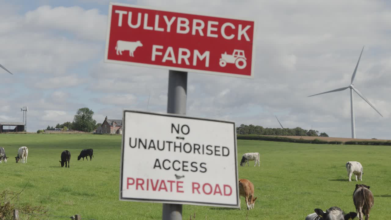 Camera pans across Tullybreck Farm sign, revealing grazing cattle, wind turbines, and green fields under bright daylight in rural Dundee, Scotland