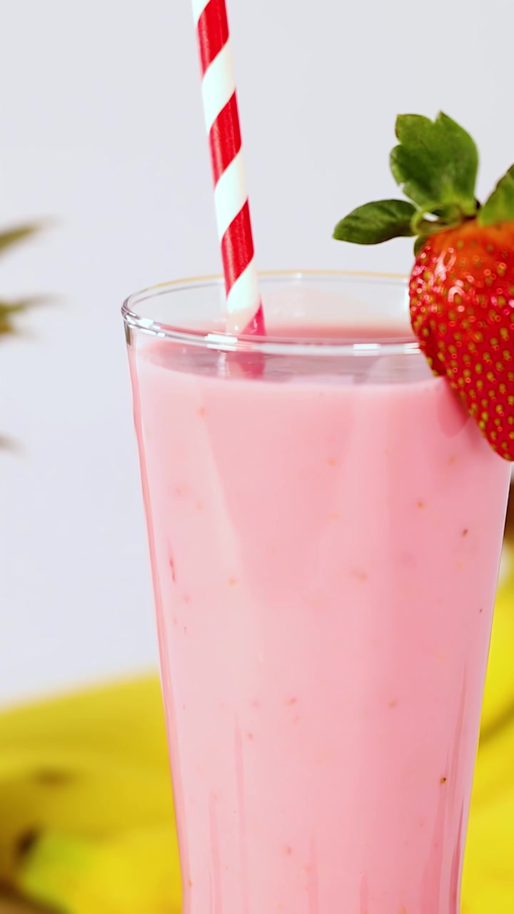 Frullato di fragole rosa in un bicchiere alto, guarnito con fragola, illuminazione brillante, sfondo di frutta tropicale.