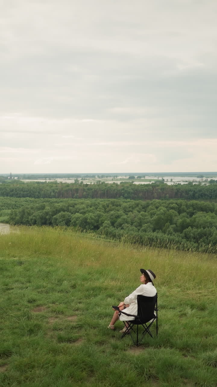 A woman in a hat, sitting comfortably on a chair in a grassy field, while an artist paints her near a serene lake. The expansive landscape, with its peaceful atmosphere and cloudy sky