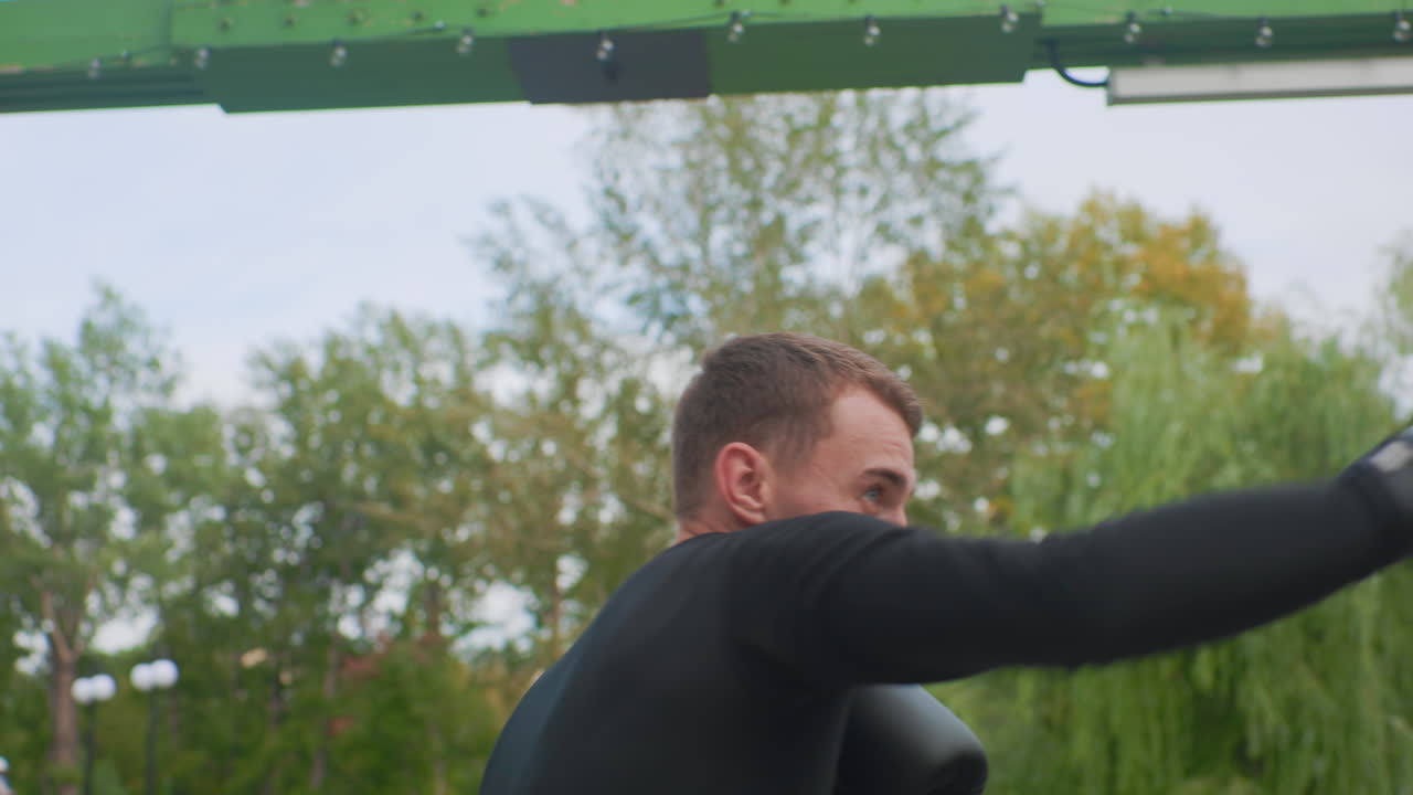 Focused caucasian boxer training outdoors, closeup guard with black gloves, intense gaze and compact stance, rhythmic jabs and defensive drills, green park background with trees and metal frame