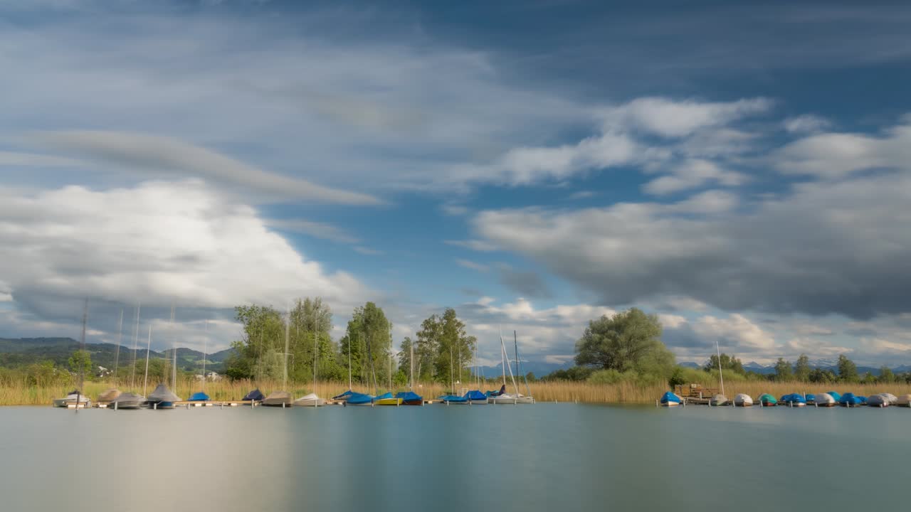 Sailboats anchored near lakeshore edge by reeds bob and sway as clouds rush over in sky