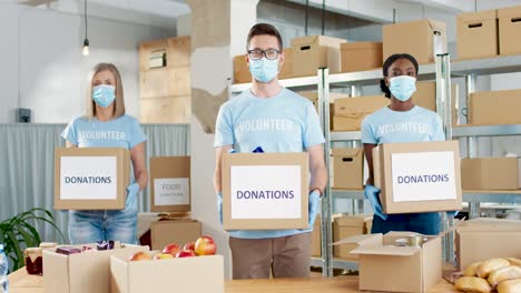 Grupo-Multiétnico-De-Voluntarios-Con-Máscara-Facial-Sosteniendo-Cajas-De-Donación-Y-Mirando-A-La-Cámara-En-Un-Almacén-De-Caridad