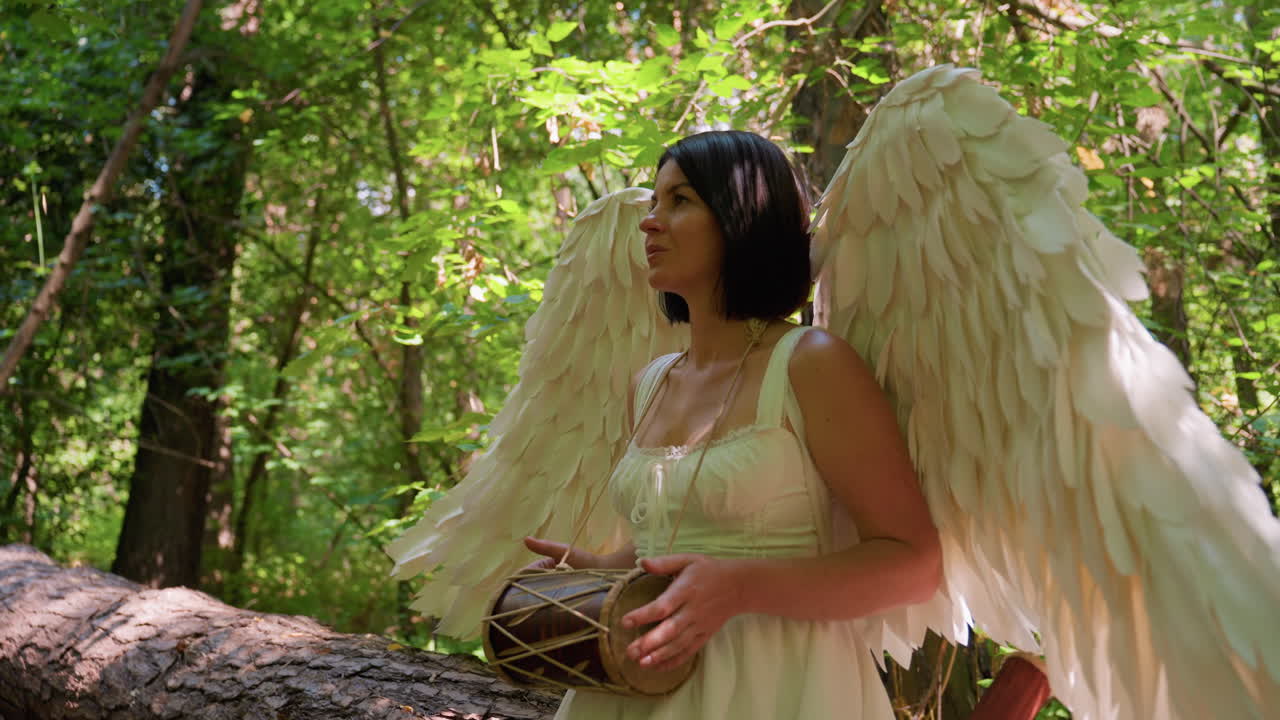 Woman dressed as angel with large white wings stands in forest holding small drum, sunlight illuminating feathers and green leaves, creating serene mystical atmosphere of harmony
