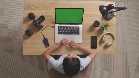 top view of asian woman video editor thinking about something while using green screen laptop and smartphone next to the camera in the workspace at home