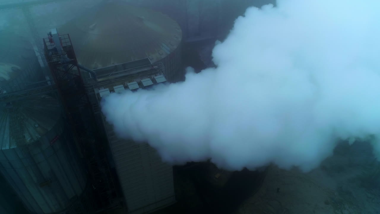High premises on the territory of grain elevator plant with pipes producing thick smoke. Grey foggy day backdrop.