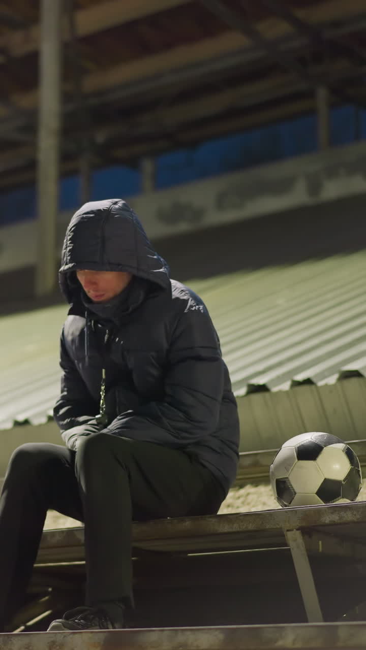 A man dressed in a black outfit and jacket sits quietly and alone in a dimly lit stadium, his hands clasped between his legs, with a soccer ball nearby