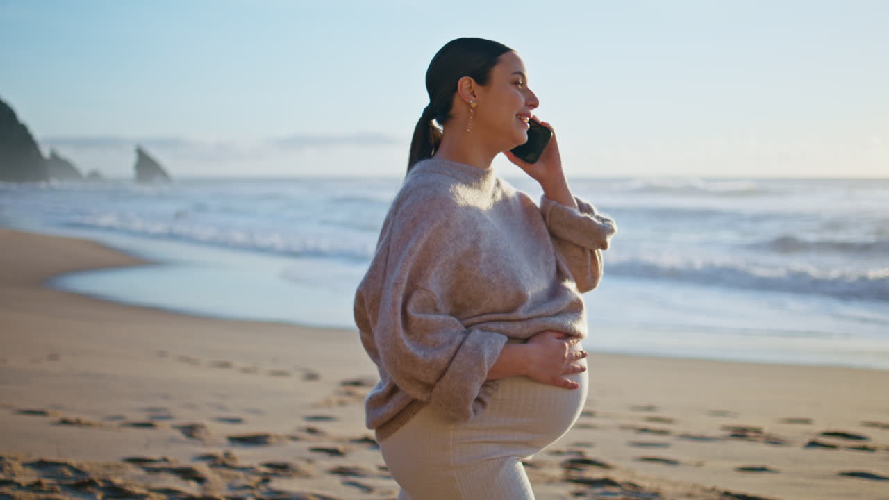 임신한 여성이 해변에서 드폰으로 전화하고 큰 배를 만지고 있습니다. 소녀가 이야기합니다.