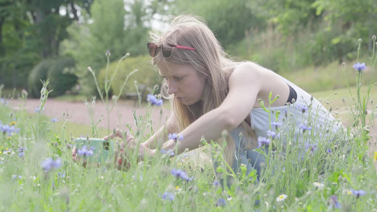젊은 금발 여성이 잔디에 무 ⁇ 을  ⁇ 고 휴대폰을 사용하여 사진을 찍습니다.