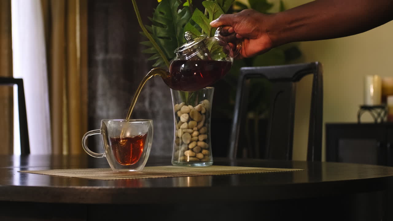 아프리카계 미국인 남자가 편안한 식탁에 앉아 있는 심장 모양의 투명한 유리 차 컵에 는 뜨거운 차를 부어넣는 중간