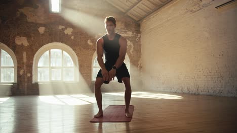 a young man in a black sports uniform in the sunny hall is engaged in fitness on a special rug. squats and other exercises for