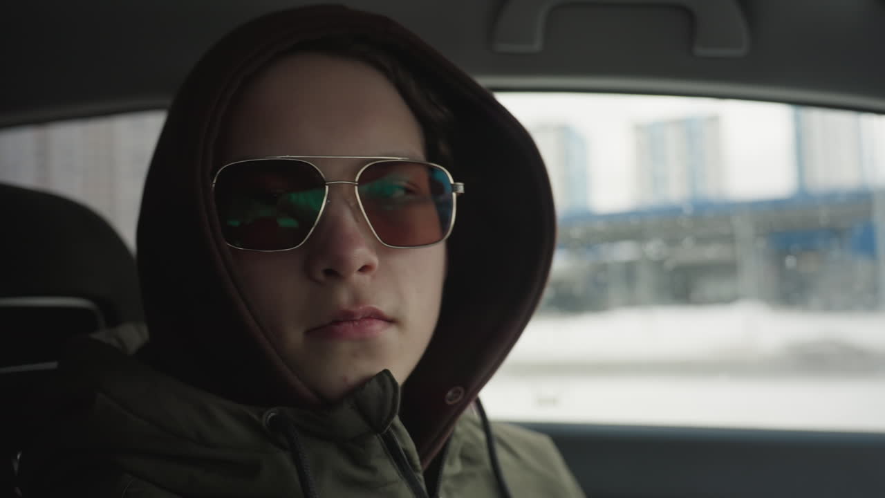 confident man in hoodie and sunglasses sitting inside car with serious expression, snowy city buildings seen through car window in background during cold winter day with soft natural light