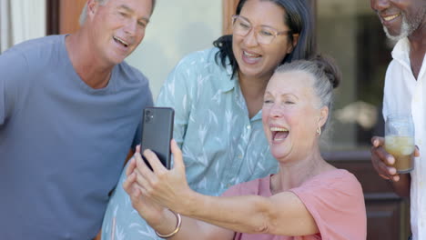 senior woman laughing with diverse senior friends while looking at smartphone in garden