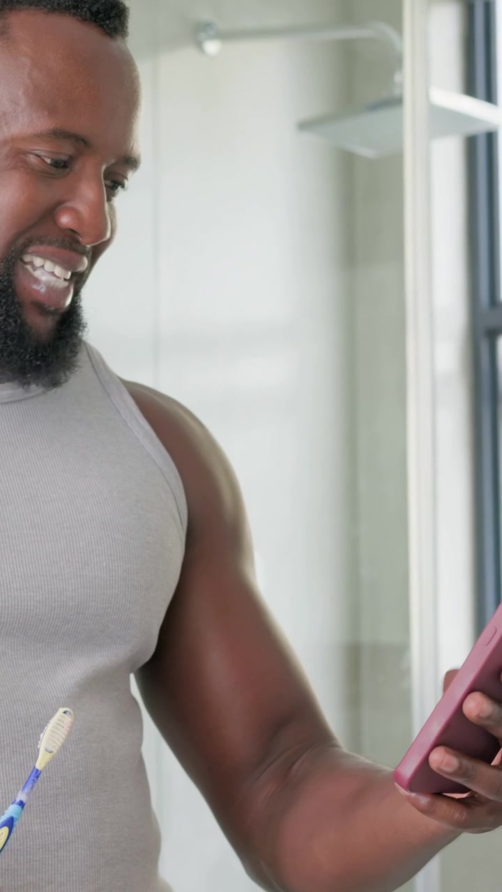 Vertical video: Brushing man in grey tank reading texts at home holding toothbrush and pink phone