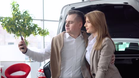 happy couple taking a selfie at a car dealership with a new car in the background