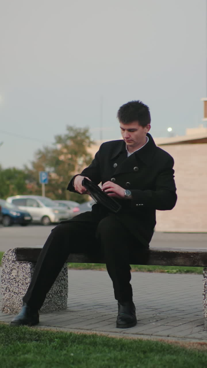 hombre de negocios en traje negro camina hasta el banco, abre el maletín en un entorno al aire libre con edificios modernos y coches estacionados en el fondo, profesional, urbano, trabajador de oficina tomando un descanso