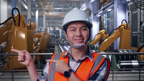 close up of asian male engineer with safety helmet smiling and pointing to side while standing with industrial robotic arms working