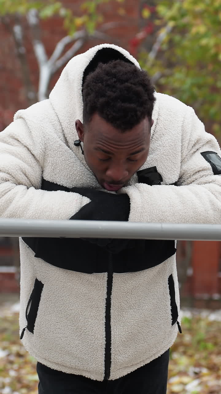 Man resting on iron bar after intense workout session in cold weather, dressed in a warm fleece jacket, he catches his breath while leaning forward, snow-covered ground and trees in background