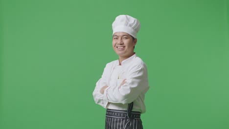 vista lateral del chef asiático sonriendo y cruzando los brazos a la cámara mientras está de pie en el estudio de fondo de la pantalla verde
