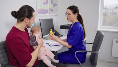 Confident-girl-doctor-in-glasses-and-blue-uniform-communicates-with-young-father-of-little-baby-girl-and-plays-with-baby-in-modern-clinic